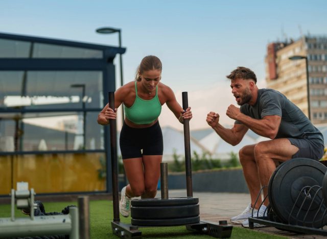 a woman does a sled workout in an outdoor gym with a trainer pushing her on