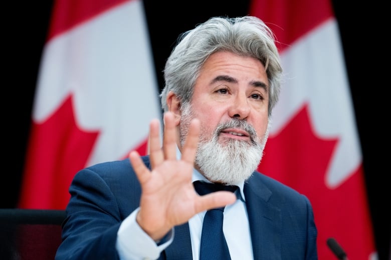 Final photo of Pablo Rodriguez, Minister of Transport. He is shown with white hair and a white beard, holding his hand up as he speaks. Canadian flags can be seen in the background. 